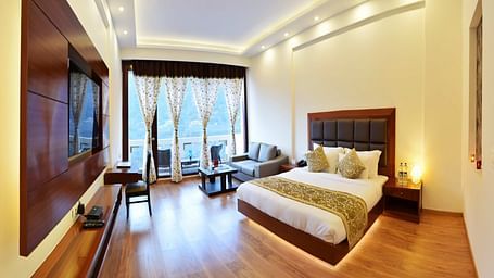 The image shows a spacious and luxurious hotel room with a king size bed, a sitting area with a sofa and coffee table, and a large window with a view.  | DLS Serenity Resort and Spa