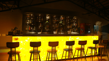 A close-up of the illuminated bar at DNC Shevaroys Resort & Spa with a row of wooden stools and shelves of bottles behind.