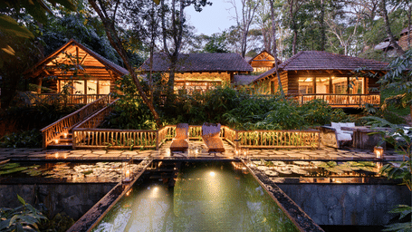 Log bungalows at Estate Plavu lit at dusk, with a stone stair, branch railings, reflecting pools with lily pads and lanterns at Earthitects Holiday Experiences.