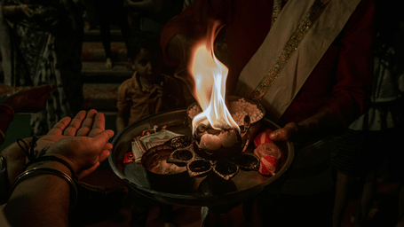A person offers a burning flame on a metal platter during a traditional religious ritual in a dim setting.