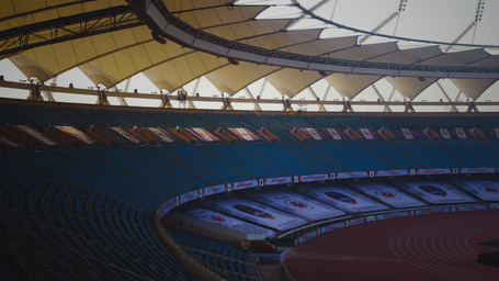 A large stadium with curved roof structure, tiered seating, and expansive arena space below