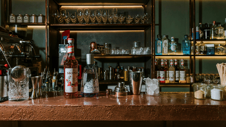 A bar counter along with a rack behind it with several glasses of different kinds along with liquor bottles.