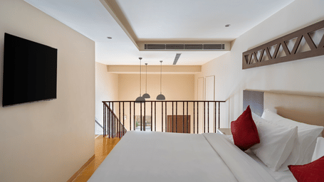 Upper-level bedroom at EsthereaRaj Leela, Ranakpur, featuring a king size bed with red pillows, a wall-mounted TV, and a railing overlooking the lower floor.