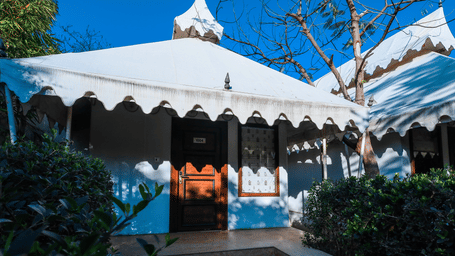 Stay near Jaipur at our cottage featuring white scalloped tent structure with a pointed roof stands under a blue sky, surrounded by green bushes and trees with bare branches at Estherea Resorts & Spa.