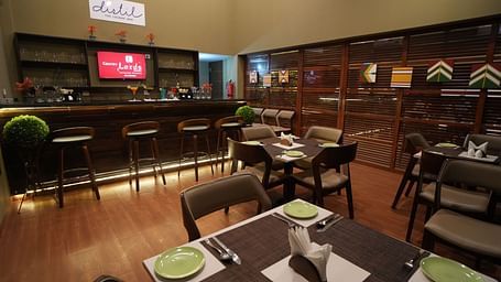 A warm, modern lounge bar with wooden flooring, neatly set dining tables, high bar stools, a well-stocked bar counter, and decor featuring colourful wall art - Gaurav Lords Resort Shrivardhan