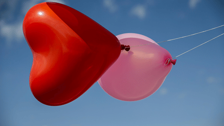 Red heart-shaped balloon and pink balloon floating together against a clear blue sky.