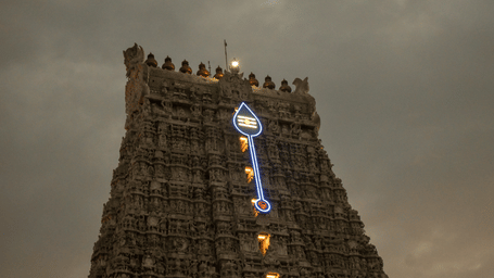 The intricately carved temple gopuram is illuminated by a digital light display under a dramatic, overcast sky.