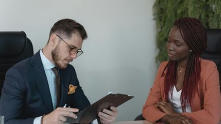 A man in a suit reviews documents while a woman in a coral blazer looks on.