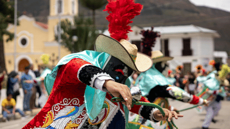 Dancers in vibrant red and teal traditional costumes with feathered hats performing in a sunny village square.