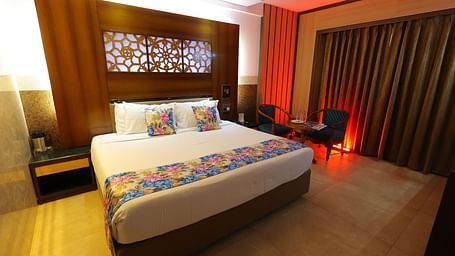  Overview image of a deluxe room at Hotel Amar with a bed, table, TV and huge window with curtains. 