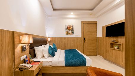 Deluxe Room at Hotel Corporate Suites featuring a plush bed facing the TV mounted on the wall and a nightstand with a telephone on it with a lamp lit over it