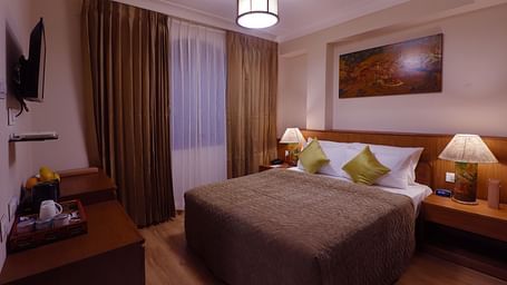 Deluxe room featuring a bed with yellow pillows, bedside tables with lamps, a mounted T.V, a cabinet, a wall art and brown and white curtains at Denzong Shangrila Lords Inn Gangtok.
