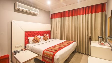 A hotel room with a plush bed featuring a patterned runner, layered curtains, a white headboard, and an AC unit, illuminated by soft lighting. - Hotel Diamond Plaza, Chandigarh.
