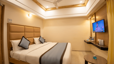 Queen bed with grey runner, padded headboard, wall-mounted Smart TV, and small round teapoi table in a guest room at Hotel Grand Sabarees Madurai.