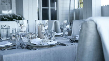 A close-up view of an indoor dining table neatly set with white tablecloth, silver cutlery, and various glasses.