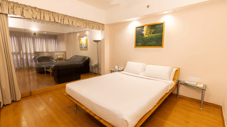 A view of the suite at Hotel Horizon, Trivandrum, featuring a queen size bed, white sheets and pillows, a wall art, and a glass partition showcasing a lounge area beyond.