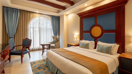 Luxurious hotel room with a large bed, blue and gold decor, a grand headboard, and arched window with sheer curtains at Hotel Hukam's Lalit Mahal.
