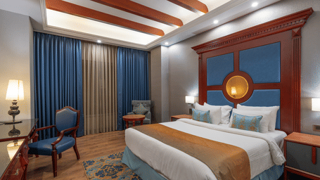 Ornate hotel bedroom with a traditional wooden headboard, layered bedding, and rich blue and gold colour scheme.