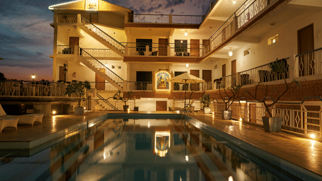 A nighttime view of Hotel Mamallaa Heritage, showcasing its illuminated facade and a beautifully lit swimming pool reflecting the lights.