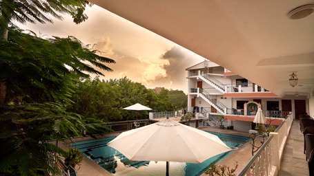 Elevated view of an outdoor resort swimming pool featuring white patio umbrellas, lush surrounding trees, and multi-story guest room balconies under a sunset sky - Hotel Mamallaa Heritage
