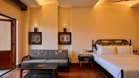 A hotel room featuring a double bed with white linens, a grey two-seater sofa, a small dark coffee table, and a wooden door on the left at Hotel Sonar Bangla, Ajodhya Hills.