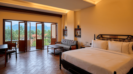 A bedroom featuring a large bed with white sheets, a small grey sofa, and open double glass doors leading to a balcony overlooking trees at Hotel Sonar Bangla, Ajodhya Hills.