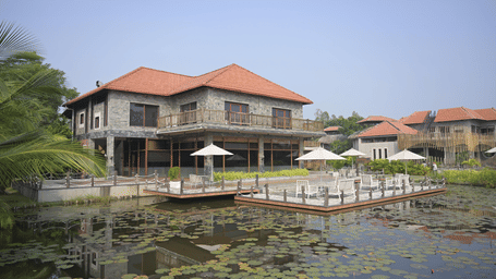 Hotel Sonar Bangla, Sundarban exterior at dusk with stone-clad building, terracotta roof, lily pond, wooden deck, patio umbrellas, and lounge seating.