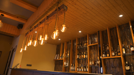 Bar counter at Hotel Sonar Bangla, Sundarban with bulbs hanging on ropes, wooden ceiling, and shelves displaying liquor bottles.