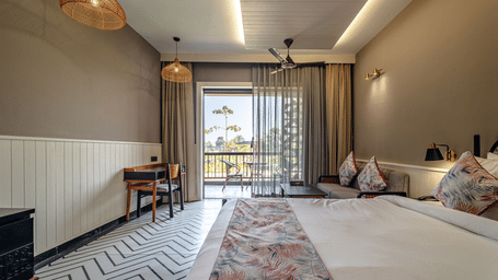A spacious deluxe bedroom with a large bed, soft lighting, stylish seating area, and balcony doors opening towards the outdoor view | Hotel Sonar Bangla, Susunia