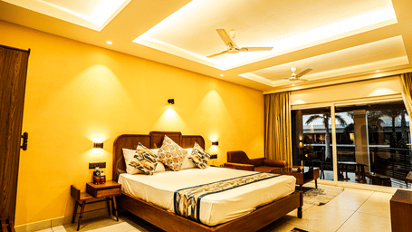 Spacious premium bedroom with a king size bed, warm yellow lighting, bedside lamps, and balcony seating area | Hotel Sonar Bangla, Tarapith