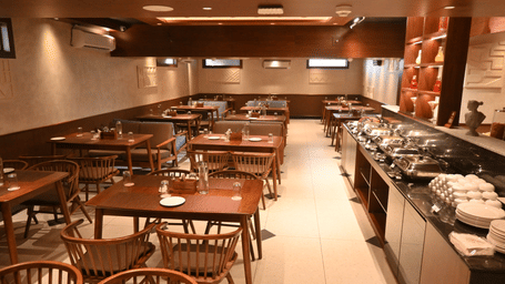 A restaurant dining room set up for a buffet, featuring rows of empty wooden tables with woven cane chairs, a long serving counter with covered chafing dishes on the right, and warm wooden panelling and recessed lighting throughout at Hotel Sonar Bangla, P