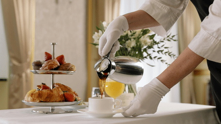 Waiter in white gloves pouring tea and serving a tiered tray of pastries.