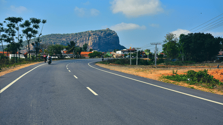 An empty two-lane motorway on a road trip from Bangalore, curving through a rural landscape towards a large rocky hill under a bright blue sky.