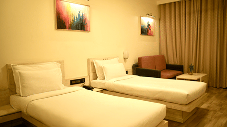 A bedroom at Infiniti Hotel, Indore featuring 2 single beds, sofa, curtains, and wall art above headboards.