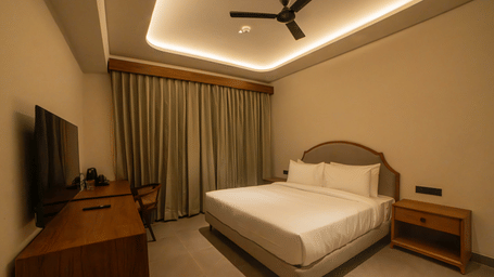 A suite featuring a king size bed in white linens, a dark wood table with a television, a ceiling fan, and long neutral curtains at Jivana Wellness Retreat, Bhopal.
