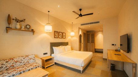 A room with a king size bed, a floral patterned sofa with a light wood coffee table, a wall shelf, and a ceiling fan in a brightly lit space at Jivana Wellness Retreat, Bhopal.
