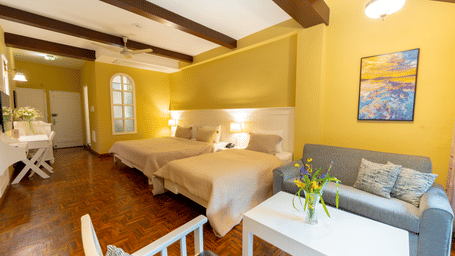 A spacious family suite featuring multiple beds, warm yellow walls, and exposed dark wood ceiling beams - Johnson Lodge & Spa, Manali