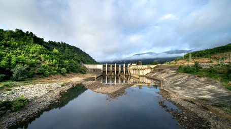 A river is seen flowing towards a dam structure with gates under a cloudy sky.