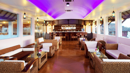 An indoor lounge bar area with wicker sofas, white cushions, and a curved bar counter under a purple-lit ceiling | Le Pondy