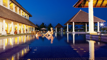 An outdoor swimming pool with blue tiles illuminated at night next to lit resort buildings and pavilions | Le Pondy