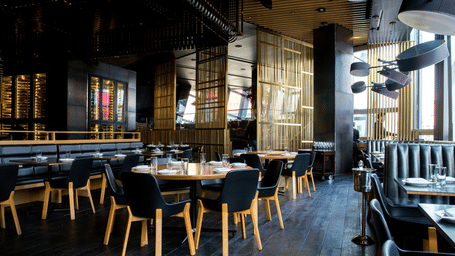 An interior view of a modern restaurant with dark flooring, wooden tables, and dark blue chairs arranged neatly.