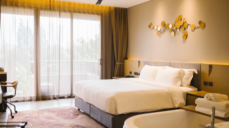 A bright and cosy room featuring a double bed, stylish decor, large windows with balcony access, ceiling fan, and modern furnishings.