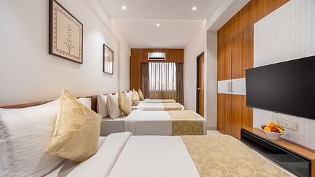 A hotel room contains four neatly made single beds, a mounted television, and simple framed artwork on the walls - Lords Eco Inn Ambaji.