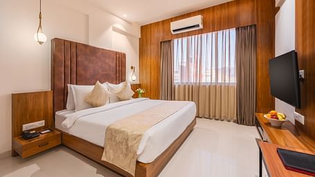 A comfortable king-size bed with soft pillows and a decorative throw faces a mounted television in a well-lit hotel room with a window - Lords Eco Inn Ambaji.
