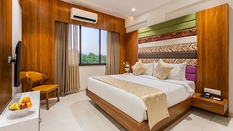 A comfortable king-size bed with plush pillows and a throw dominates a hotel room with a TV and a cushioned chair - Lords Eco Inn Ambaji.
