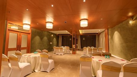 A warmly lit banquet hall at Lords Eco Inn Apple Gate is set with round tables draped in white, surrounded by chairs with gold sashes.