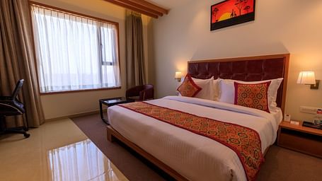 A well-lit hotel room at Lords Eco Inn Apple Gate features a double bed with patterned accents, a desk, a chair, and window curtains.