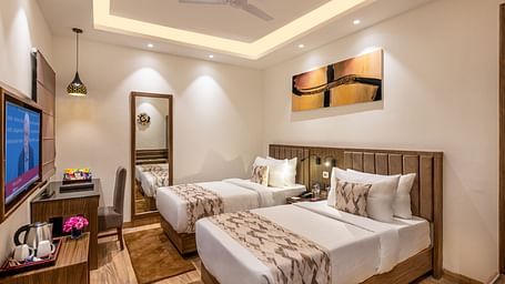 A hotel room with two twin beds, a flat-screen TV, and a work desk. It features a unique tray ceiling with lighting, a ceiling fan, and colourful abstract artwork - Lords Eco Inn Dehradun.