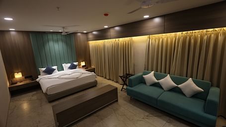 The image shows a comfortable hotel suite with a bed, a sofa with throw pillows, bedside lighting, and window coverings - Lords Eco Inn Navsari.