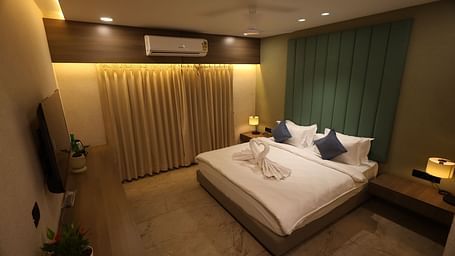 An elegant room with a king size bed featuring swan-shaped towels and pillows, bedside tables and lamps, a T.V. above an elongated desk with potted plants and illuminated draped curtains at Lords Eco Inn Navsari.
